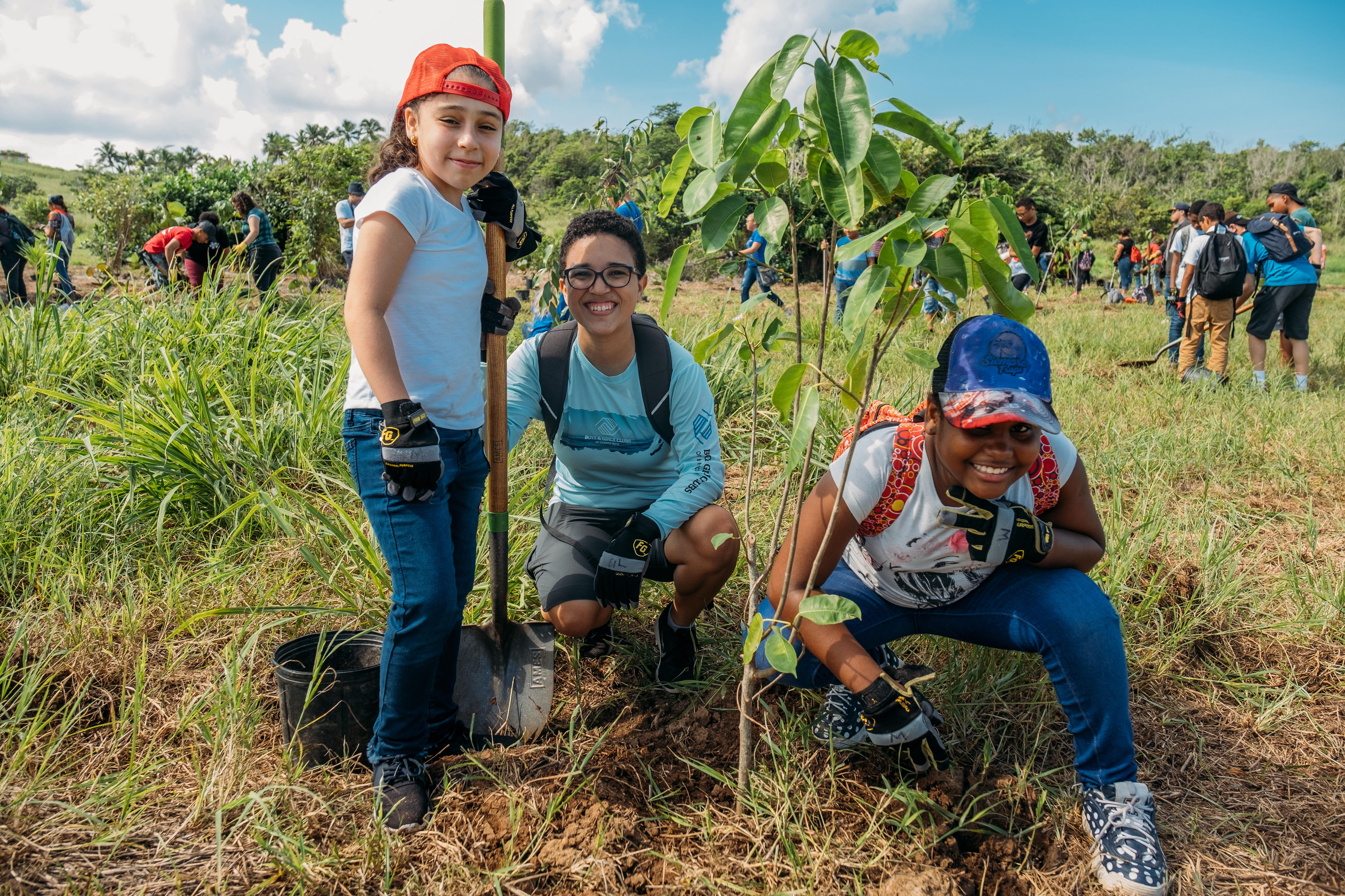 Three young people smile at the camera standing near a freshly planted tree